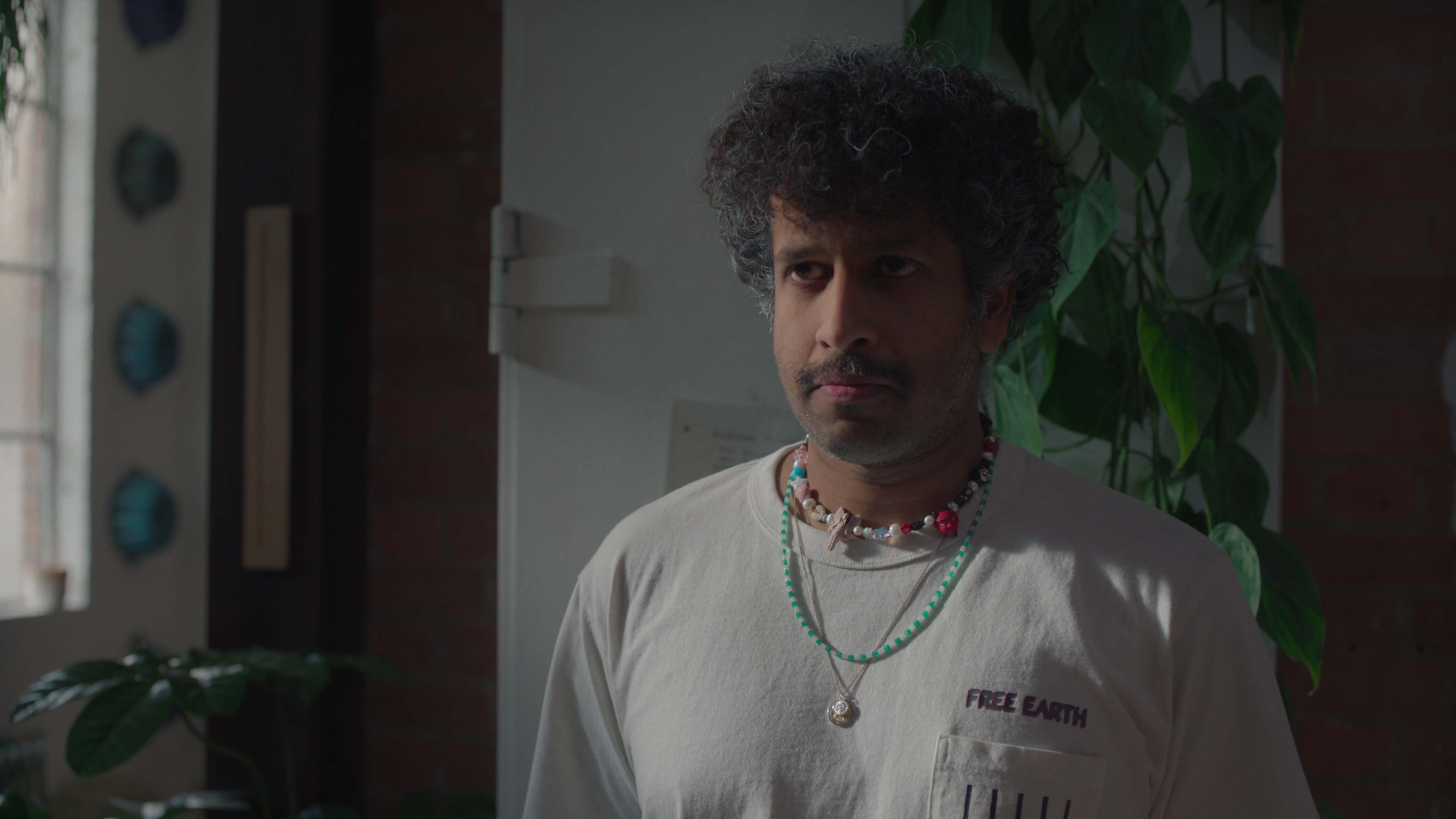 Prasanna Puwanarajah as Auggie wearing a t-shirt and jewelry in his apartment in too much