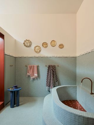 A duck egg blue, tiled bathroom with a custom, crescent bath along the right wall, with bronze tap. Along the far wall, a dado rail at half heigh with cream walls and plate artwork. Also on the back wall hangs two towel rails and a small side table to the left of the space, in front of the cupboards.