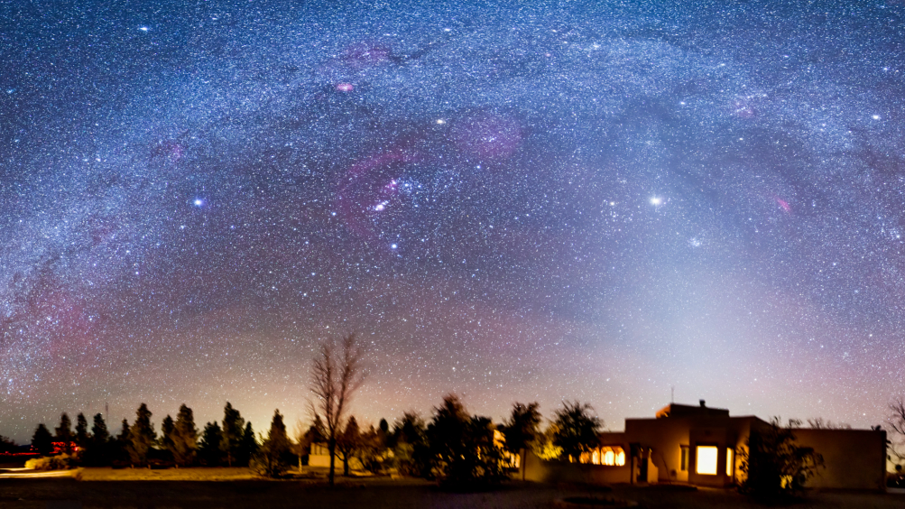 The Milky Way is pictured shining in a starry night sky above a house illuminated with artificial light. Trees can be seen lining the horizon.