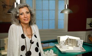 Carme Pinós in a room with mini architectural structural objects on a green table and silver pendants ceiling lights hanging from the ceiling