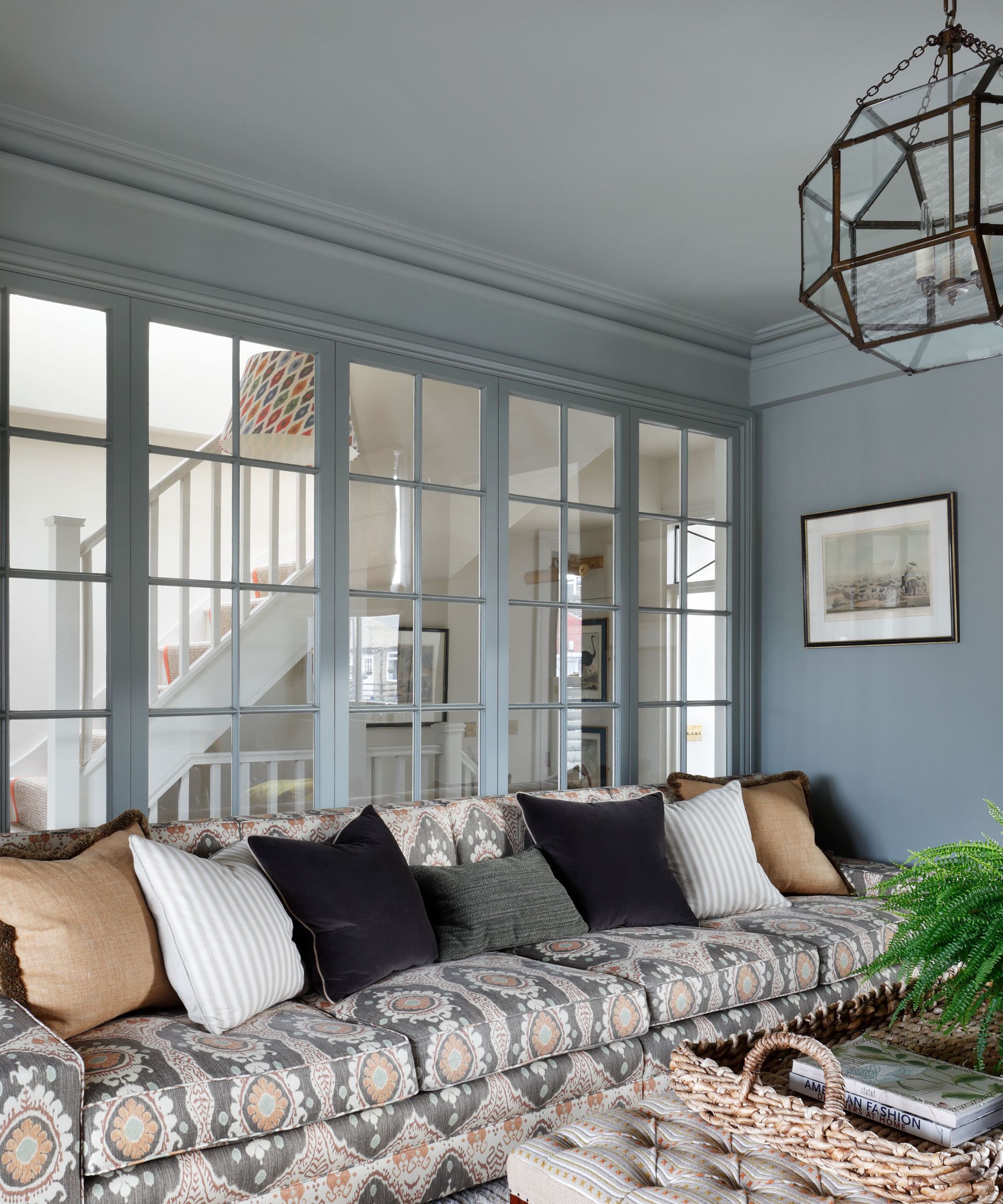 An example of broken plan living room ideas showing a grey and orange patterned sofa with black, white and orange cushions in front of a large internal window