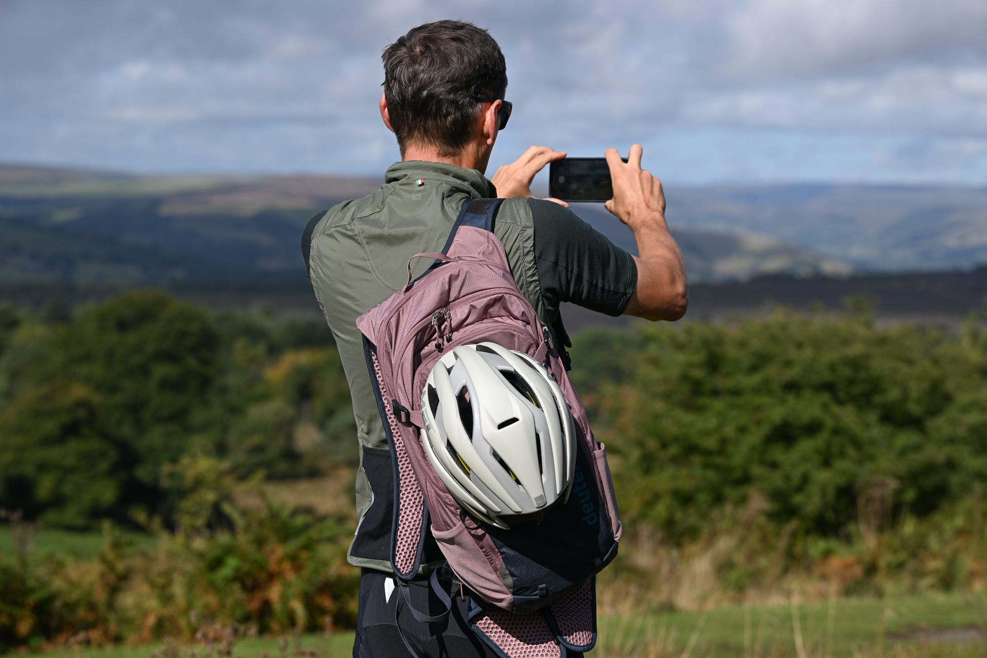 Man wearing a green jersey, black shorts and a green gilet with a pink and blue backpack on, taking a picture with his phone