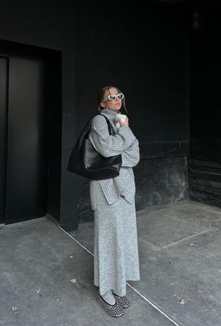 A winter travel outfit is shown in a photo of a woman standing outside wearing a gray turtleneck sweater with a matching maxi knit skirt styled with matching gray socks, embellished ballet flats, a black hobo bag, and white sunglasses