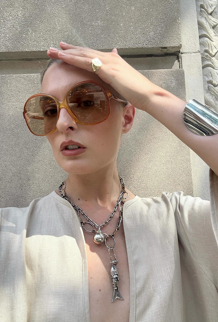 The coastal boho trend is shown in a close selfie photo of a woman wearing orange oval oversized sunglasses styled with a linen low scoop neck tan maxi dress, a chunky silver cuff bracelet, and a chunky silver lariat necklace with a fish pendant