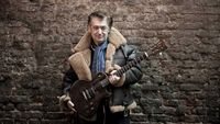 Chris Spedding holding a guitar, pictured standing against a brick wall