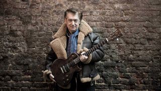 Chris Spedding holding a guitar, pictured standing against a brick wall