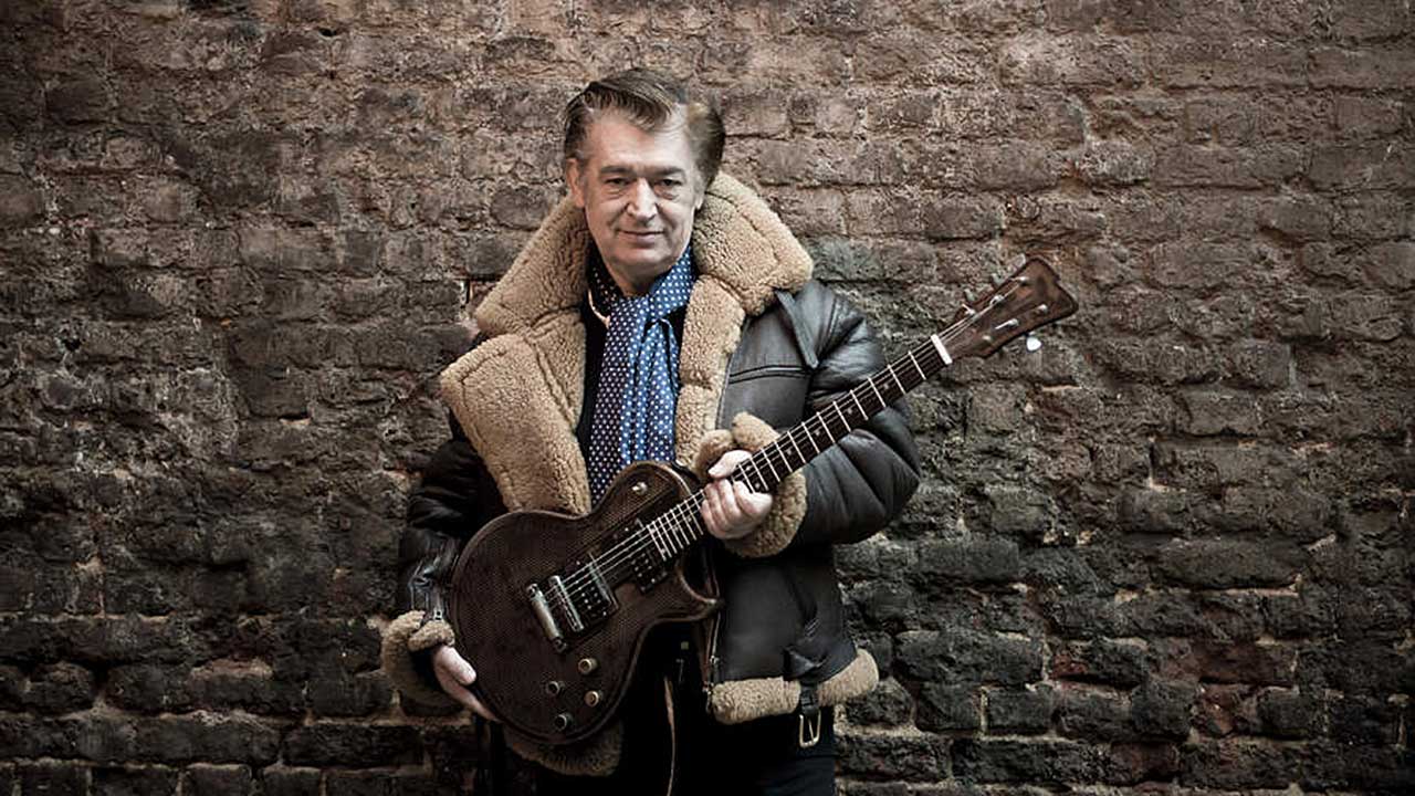 Chris Spedding holding a guitar, pictured standing against a brick wall