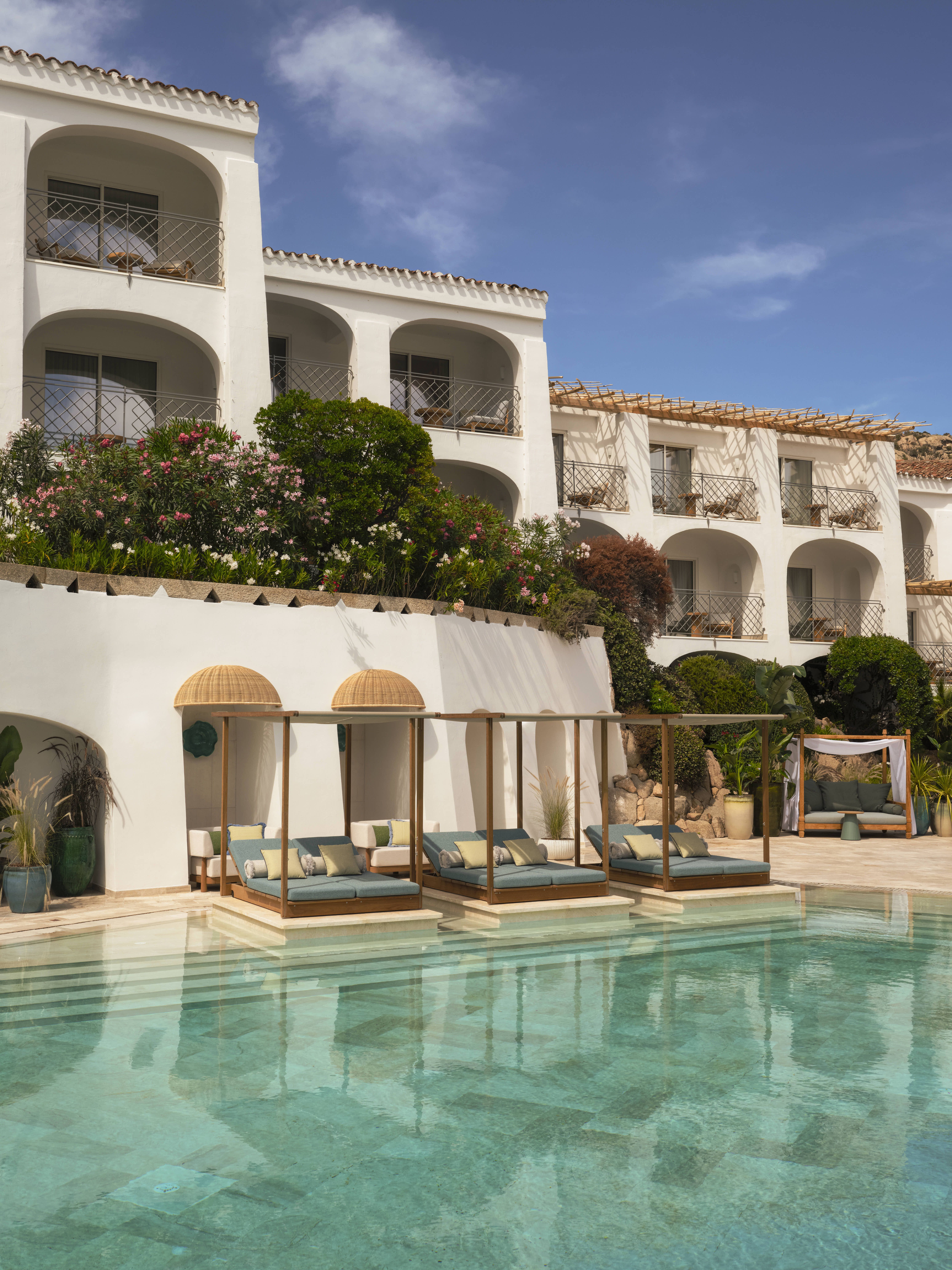 A seaside resort features spectacular, arched ceilings painted in a creamy tone, rattan and wooden furniture in playful silhouettes, turquoise accents, and a panoramic pool.