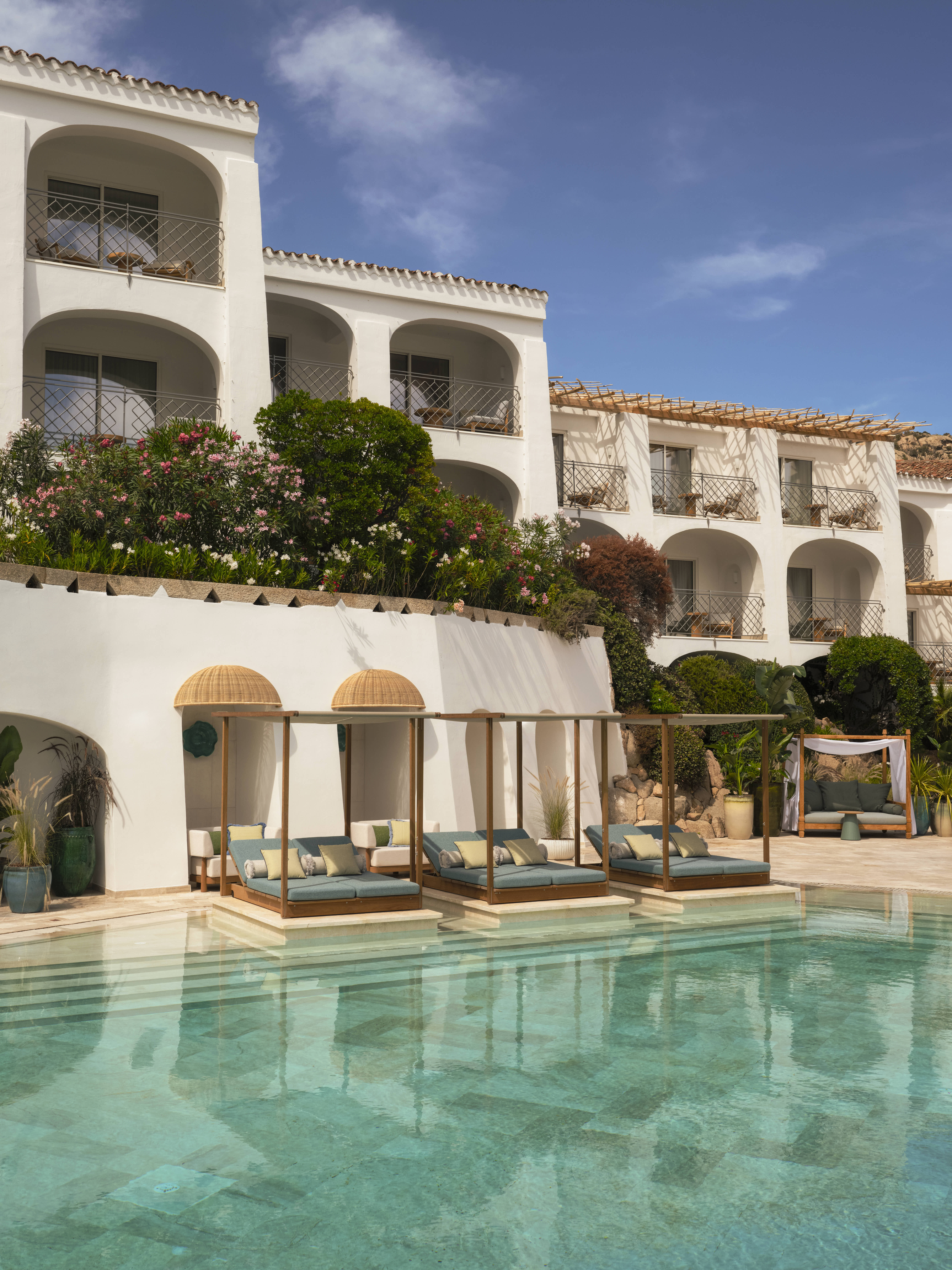 A seaside resort features spectacular, arched ceilings painted in a creamy tone, rattan and wooden furniture in playful silhouettes, turquoise accents, and a panoramic pool.
