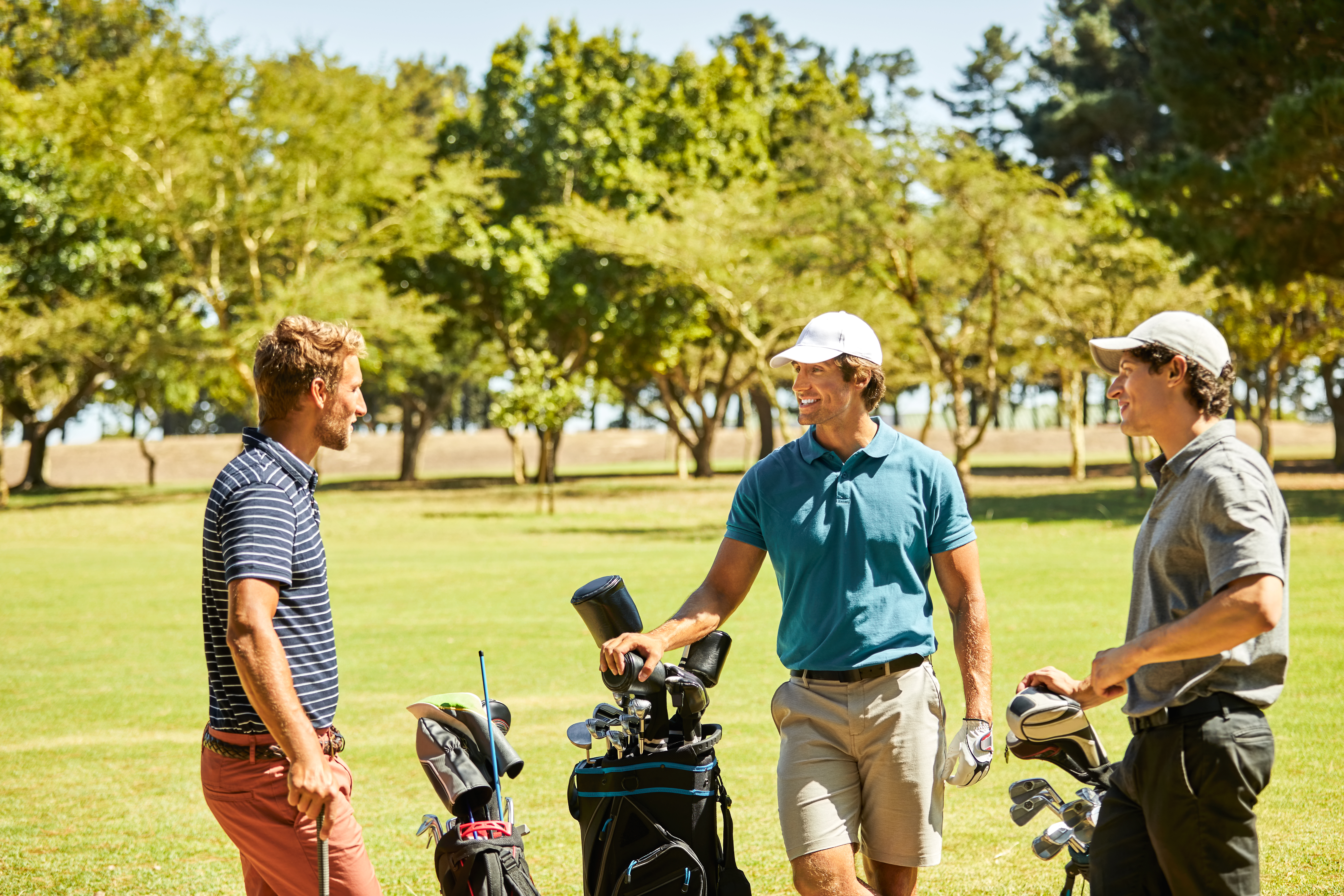 Smiling male golfers talking on golf course. Friends are standing with bags on sports field. They are spending time together in summer.
