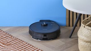 a rechargeable robot vacuum and mop in black by ultenic is photographed on a hardwood floor with a blue wall and a burnt orange terracotta and white striped rug