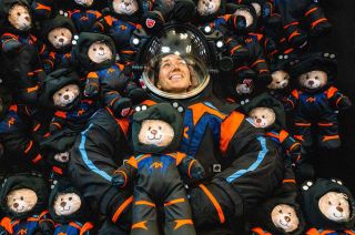 a man in a black spacesuit holds a small stuffed bear wearing a miniature version of the same suit, surrounded by dozens of other similarly clad stuffed bears.