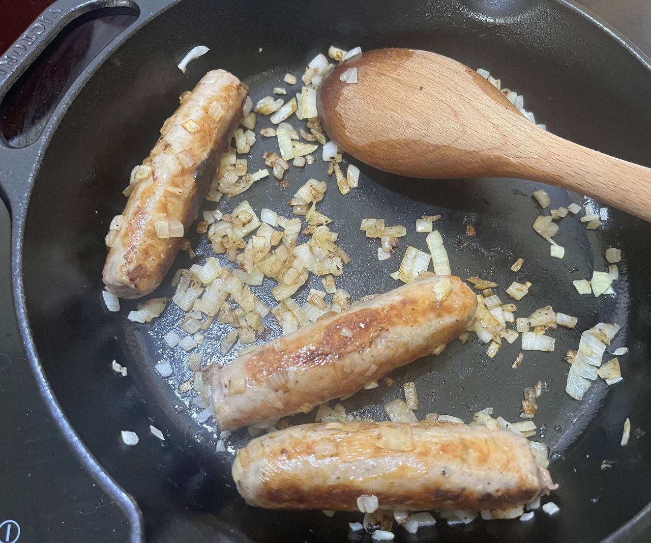 Three sausages and chopped onion in a black cast iron pan, turning golden.
