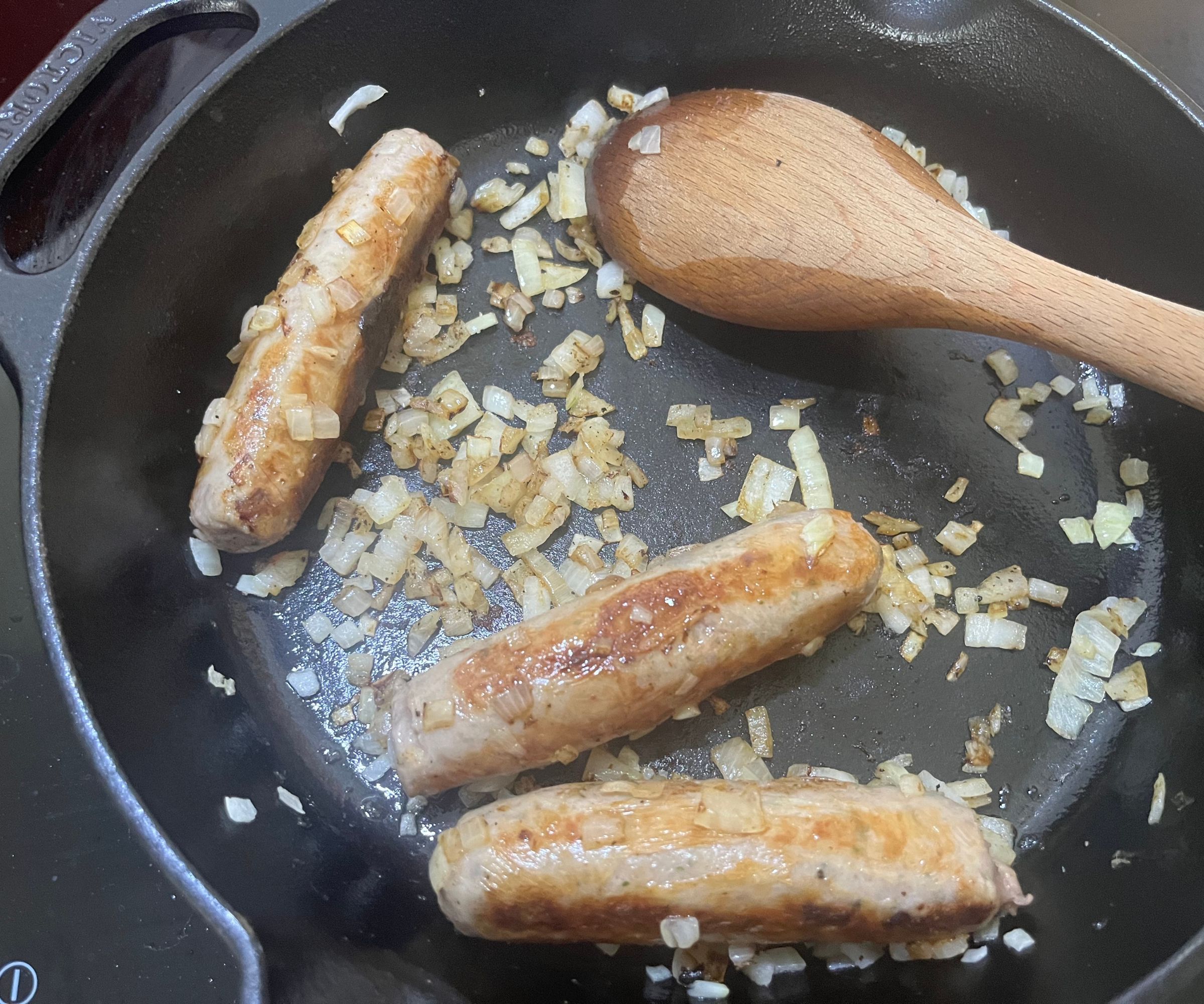 Three sausages and chopped onion in a black cast iron pan, turning golden. 