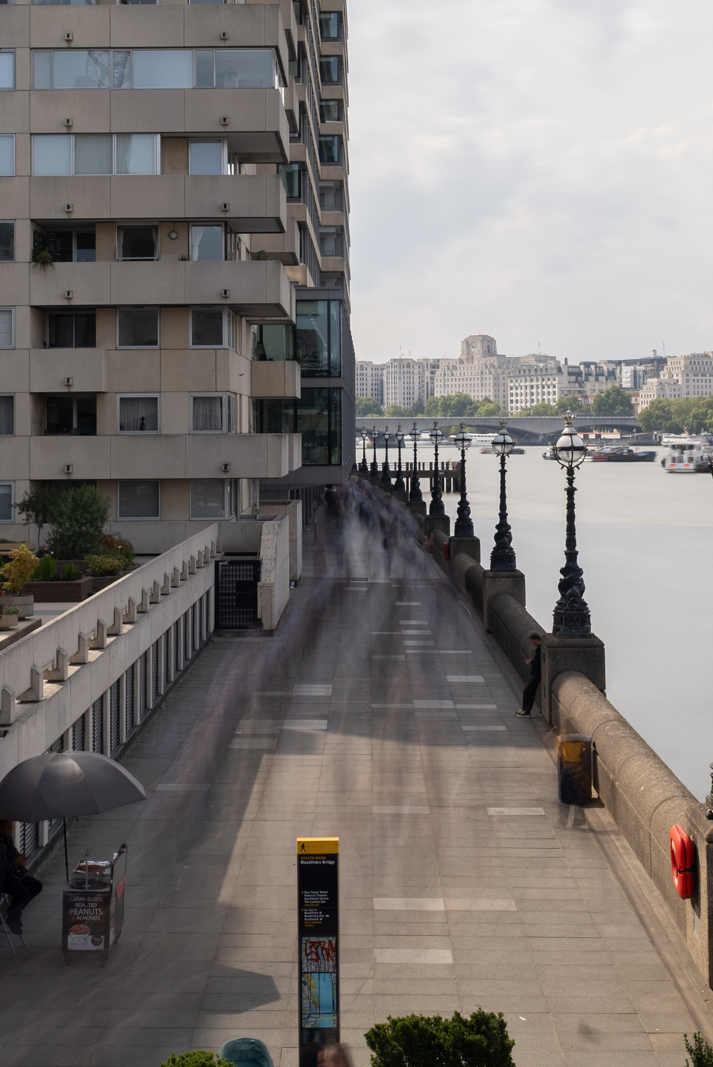 Long-exposure photograph of the South Bank in London beside the River Thames, with pedestrians blurred into ghostly streaks along the riverside walkway.
