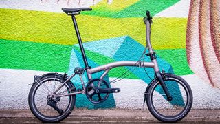 A titanium Brompton T Line with mudgaurds stands in front of a multi-coloured wall