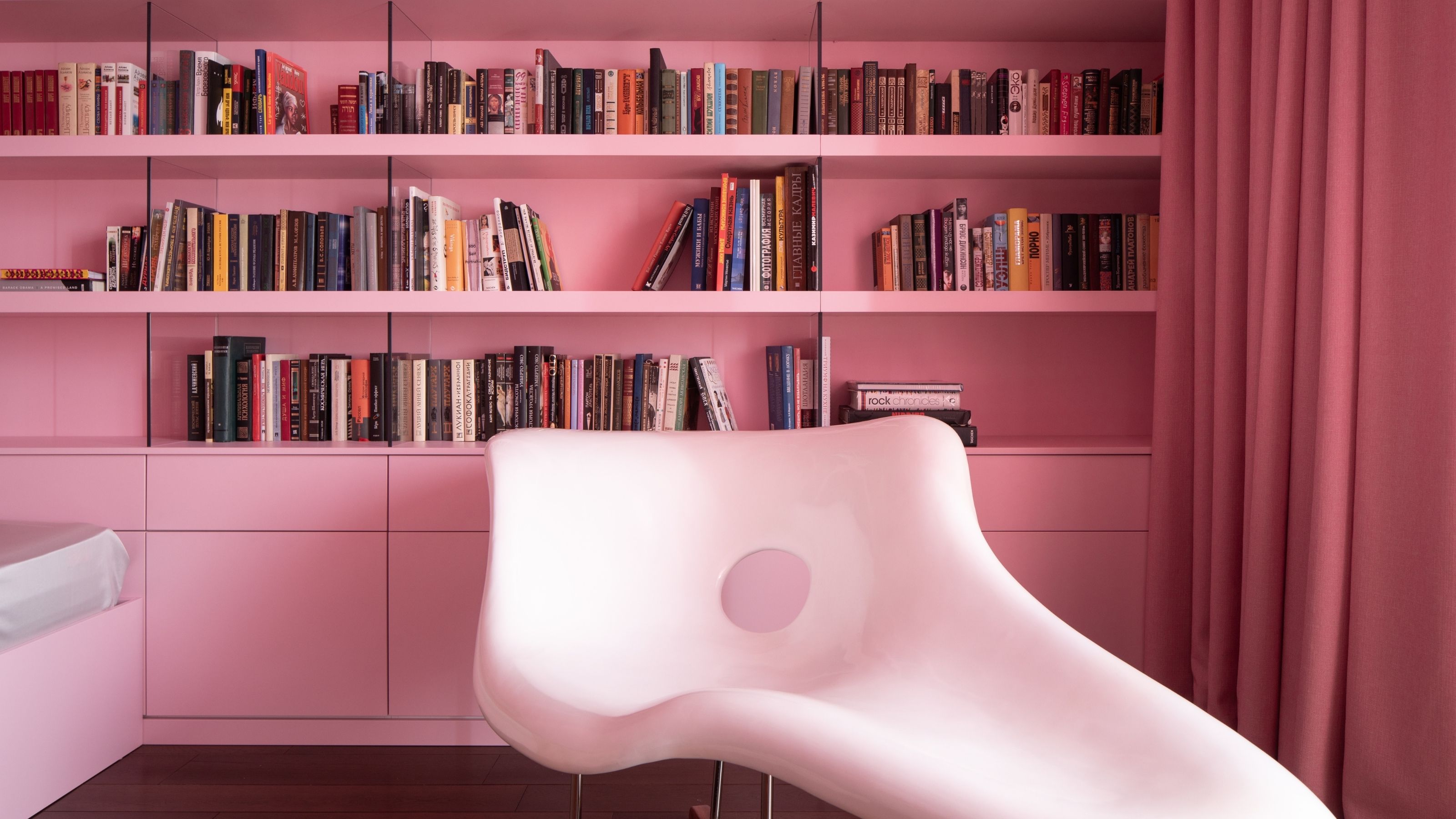 Close-up of a built-in bookshelf in a room completely drenched in bubble-gum pink. There is a modern, shiny white chaise lounge chair.
