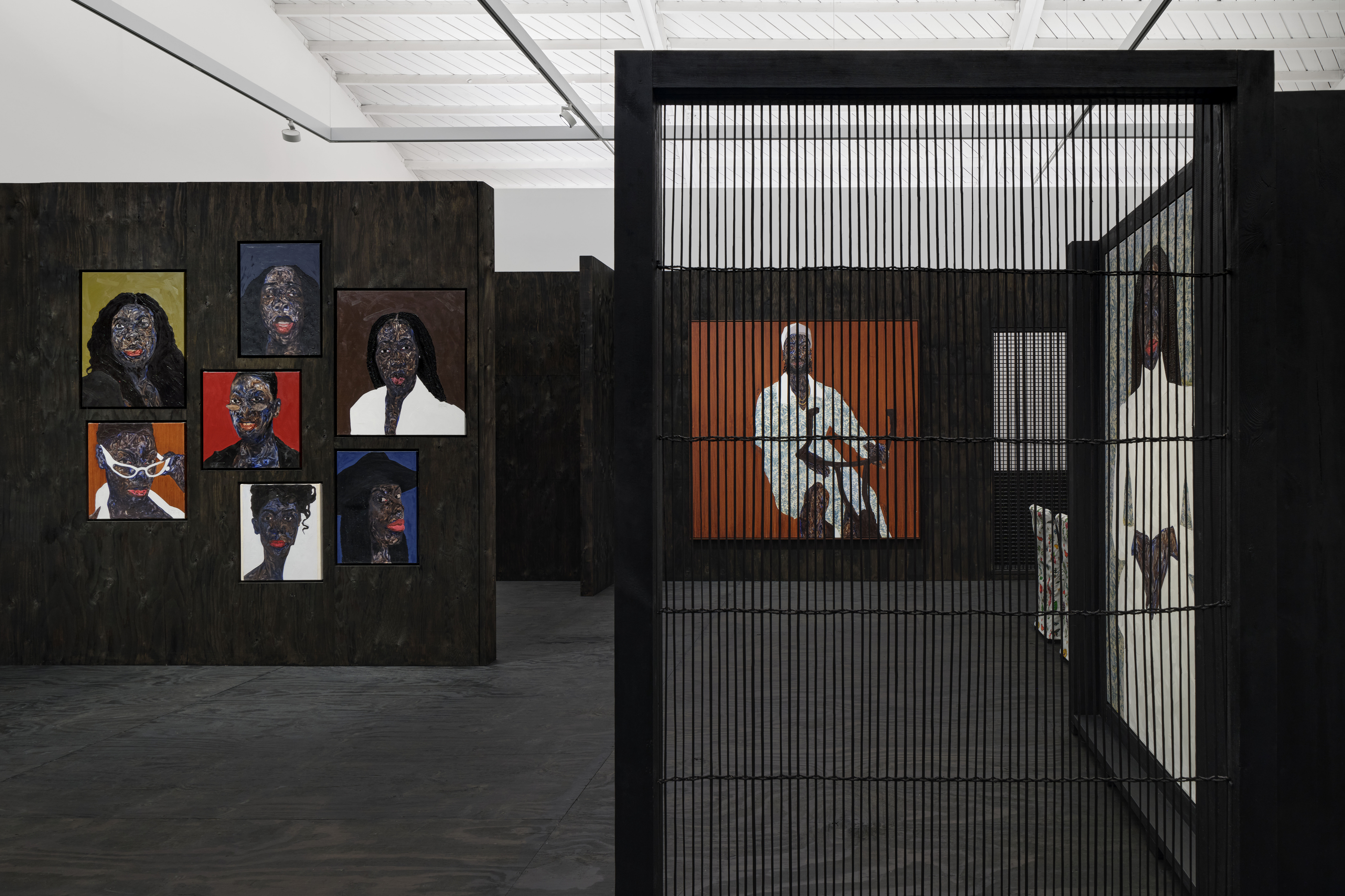 View of an art gallery installation with dark wood walls and multiple expressive portrait paintings. In the foreground, a large black metal structure with vertical bars frames additional paintings inside, while more portraits hang on the walls in the background.