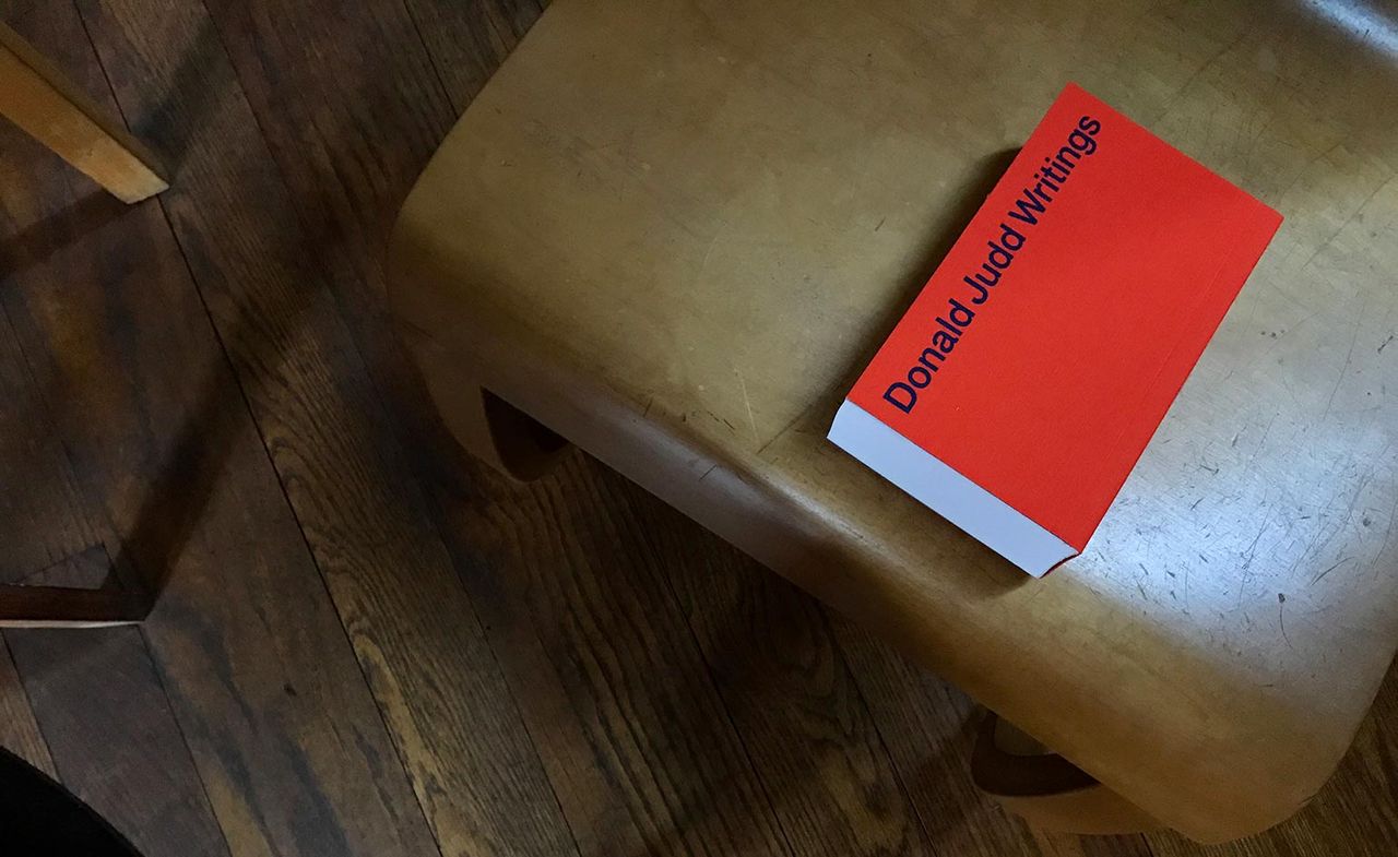 A book with a red cover says &#039;Donald Judd Writings&#039;. It&#039;s photographed on a wooden chair.