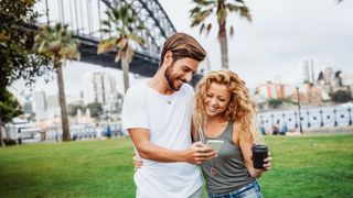 Man and woman hugging whilst looking at phone, and standing in front of Sydney Harbour Bridge