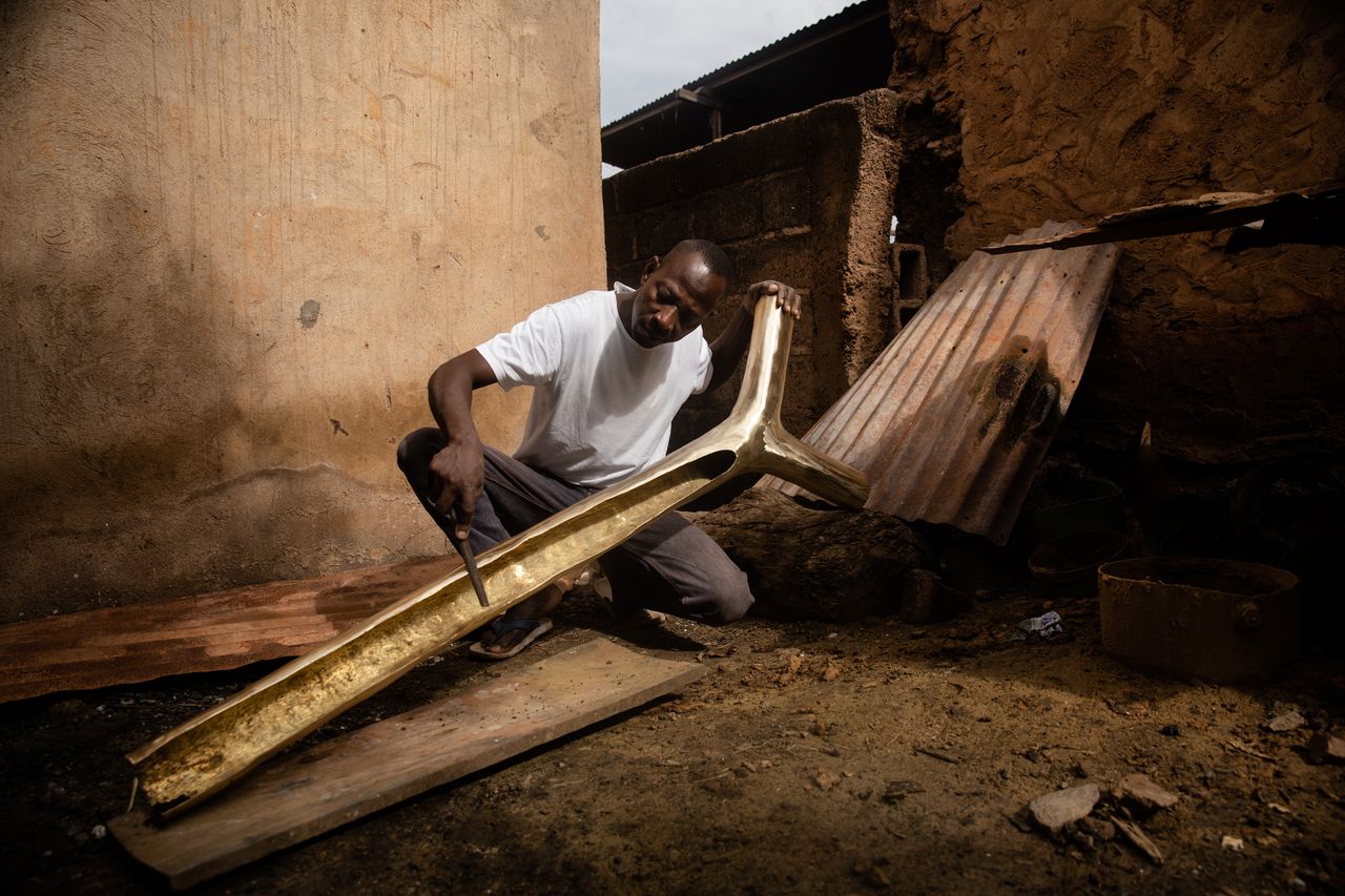 An African man crafting a long rounded object with a metal file outside.