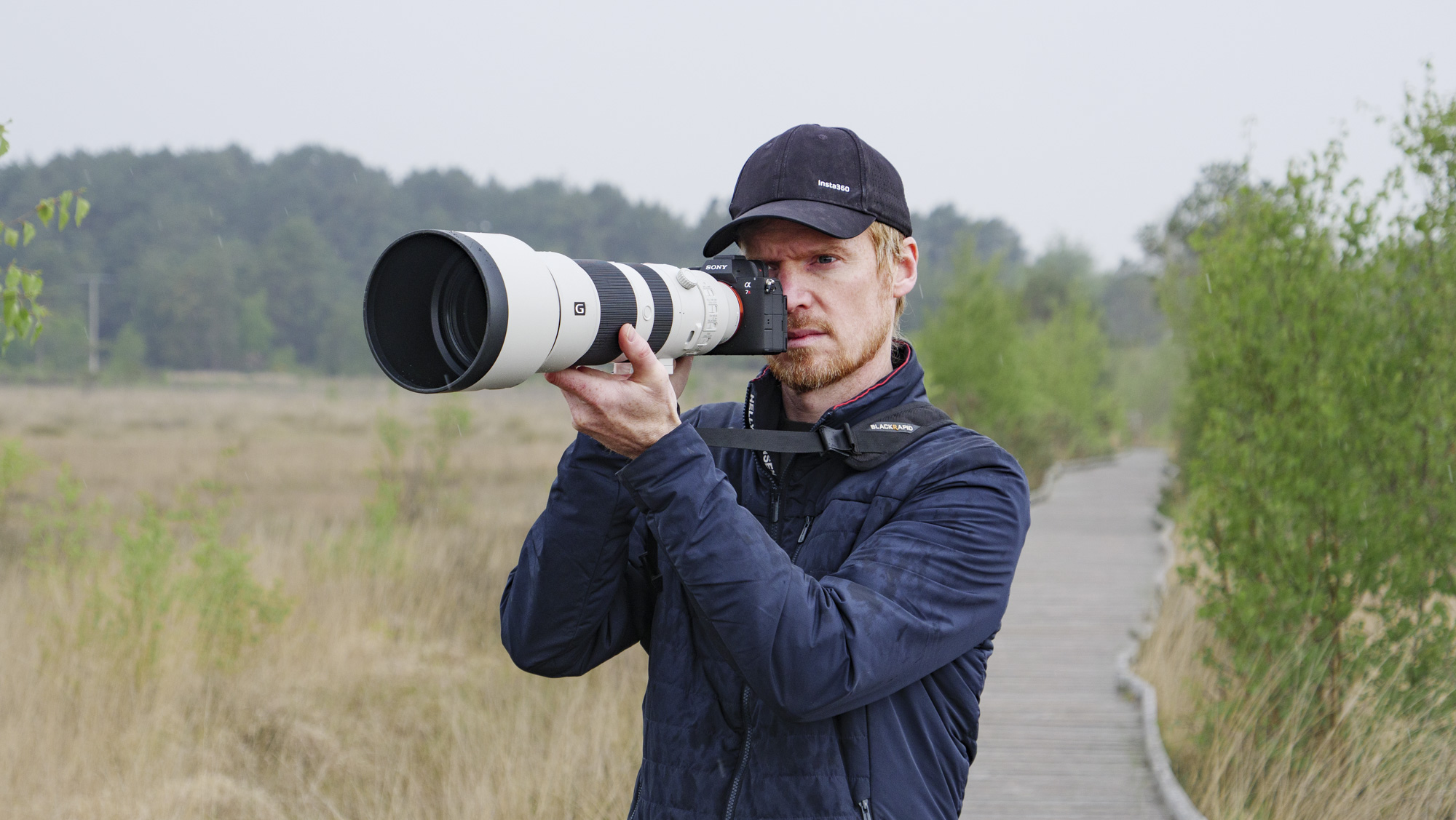 Man holding the Sony A7R VI up to his eye with the FE 400-800mm F6.3-8 G OSS supertelephoto zoom lens attached, he's standing on a boardwalk in a nature reserve
