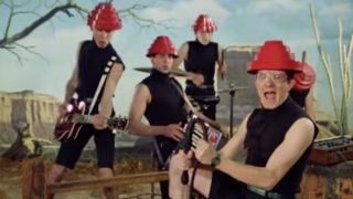 Mark Mothersbaugh in a black outfit and a red pyramid hat standing in front of the rest of Devo dressed the same in the video for "Whip It"