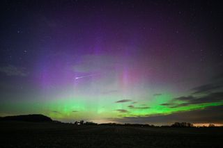 green and purple ribbons of light fill the sky and a bright white streak of a meteor is visible in the center of the image. 