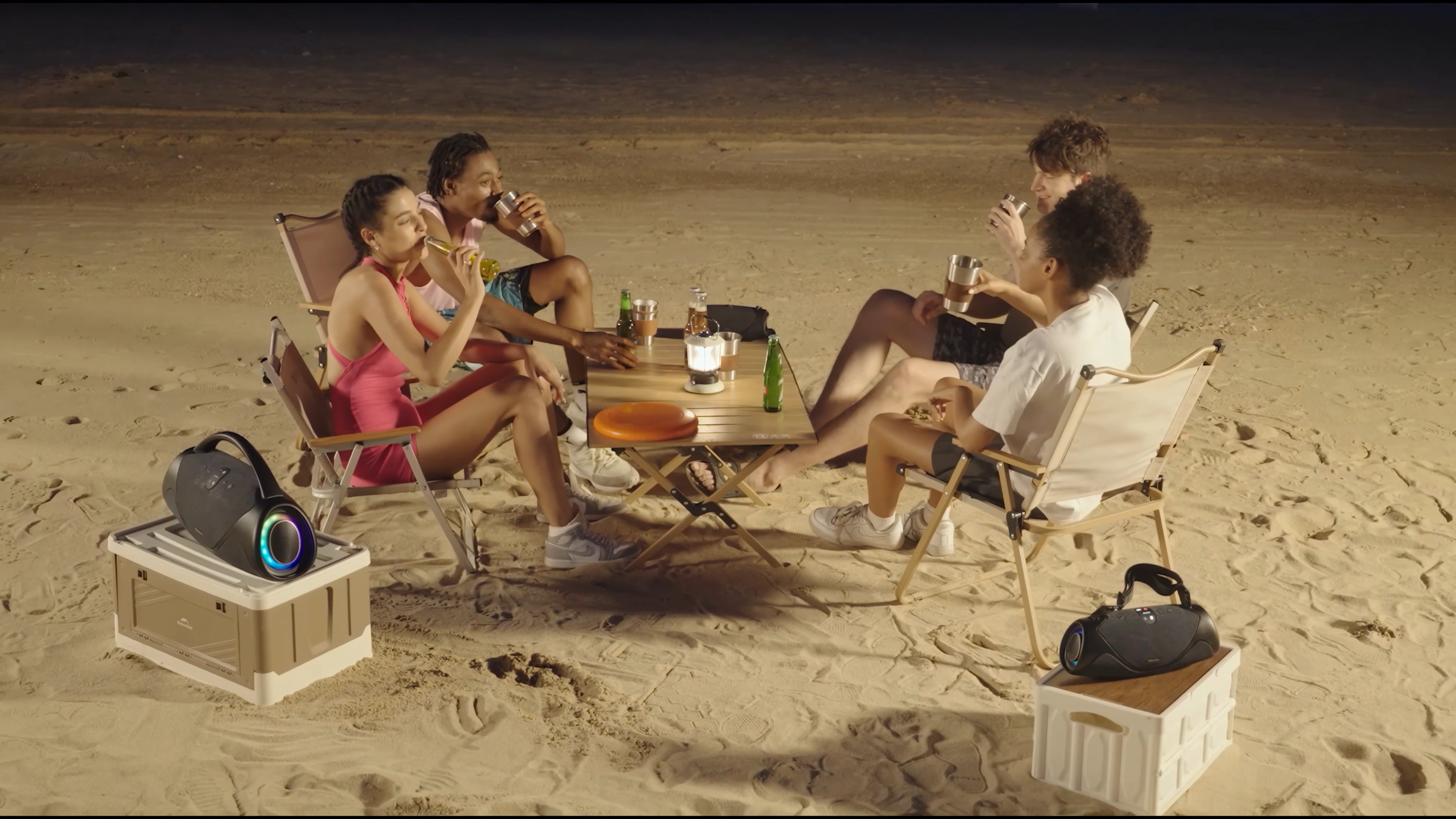 A group of friends sitting on outdoor chairs on a beach with two bluetooth speakers situated on boxes