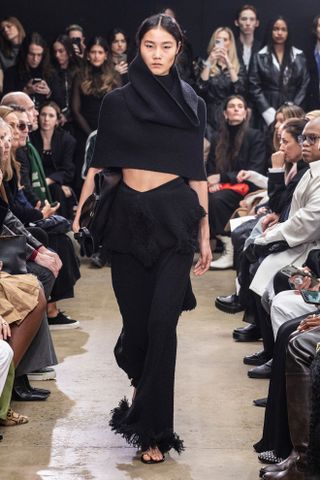 A photo of a model walking in the F/W 24 Proenza Schouler show wearing a black cape top with black pants.