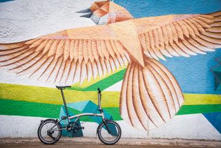 One of the best folding bikes, the titanium Brompton T-Line, leans against a wall, which has been painted with a large bird