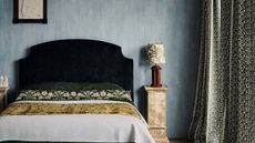 Bedroom with blue washed walls a large dark green velvet headboard, a stone plinth as a bedside table and floral drapes