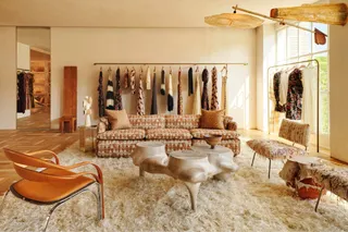 An impressively designed store features a mix of mid-century modern and modern rustic furniture in natural tones of brown, grey, and cream on a wooden floor, alongside multiple clothing rails stocked with just-as-neutral garments.