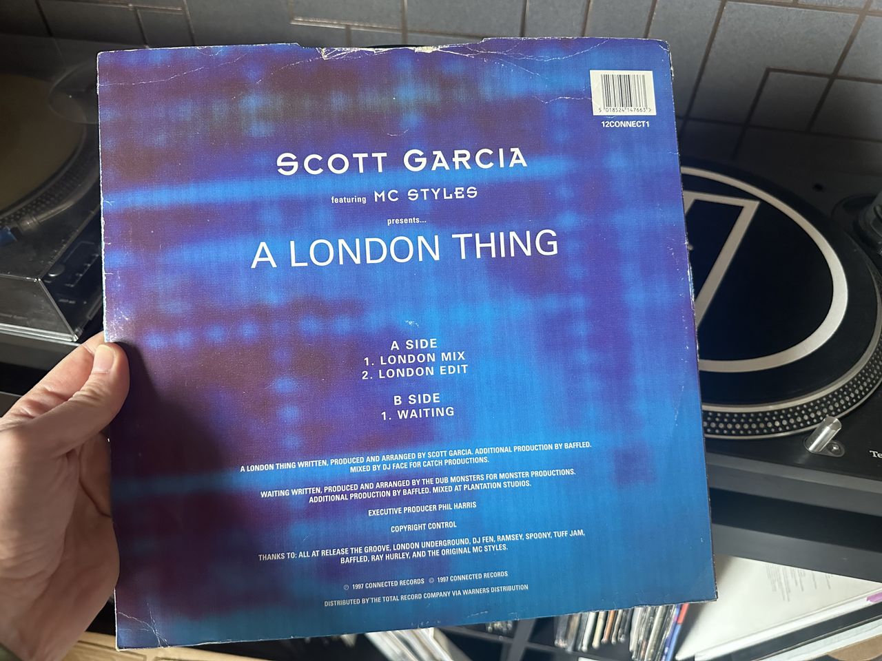 A hand holding a copy of Scott Garcia's A London Thing on vinyl. The back cover is mostly blue and lists the tracks. A turntable is visible in the background.