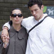A photo of Dua Lipa and Callum Turner after their London wedding ceremony walking around the city together. Dua is wearing a gray, long trench coat, white jeans, and chestnut Ugg boots.