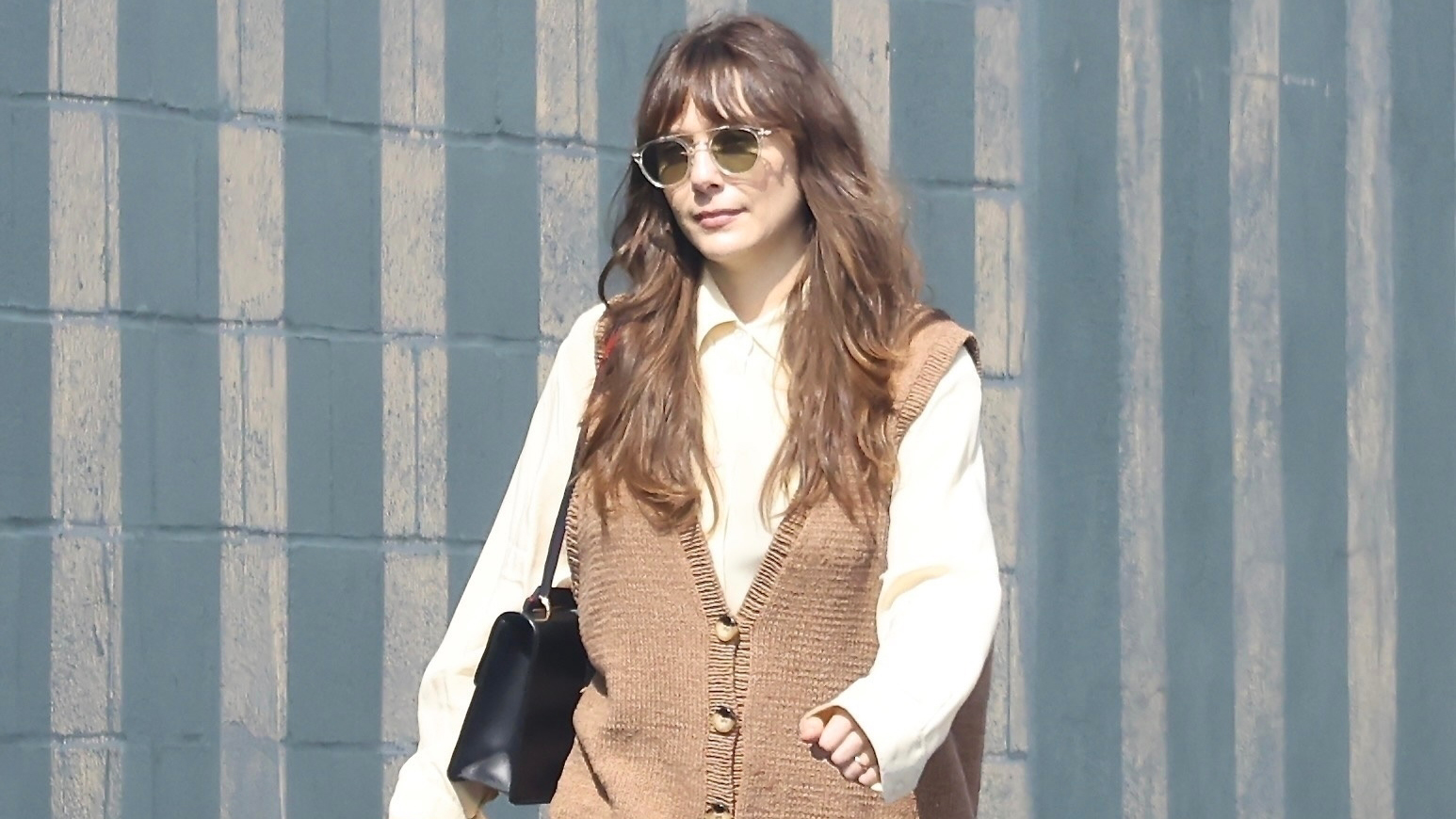 Elizabeth Olsen wearing a tan sweater vest, button-down shirt, jeans, and high-vamp pumps