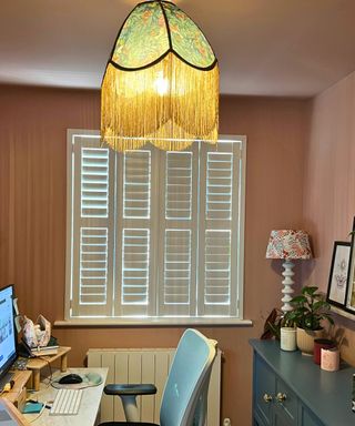 Closed white shutters with daylight showing through gaps in a terracotta home office with blue sideboard and tasselled green ceiling shade
