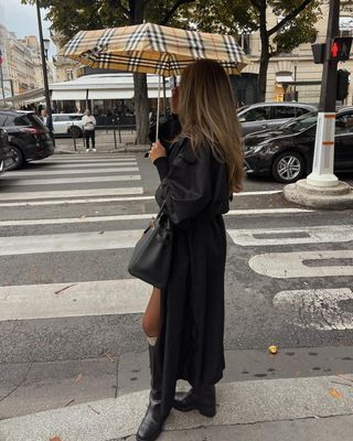 Lovia wears a black trench coat, black knee-high boots, a black bag, and a Burberry umbrella.