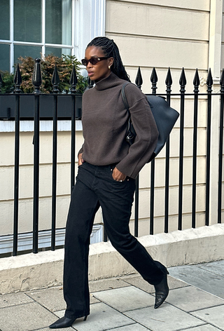 a photo of a woman walking down the sidewalk wearing a dark brown turtleneck with a black bowler bag and black jeans and black pointed ankle boots