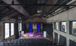Inside view of Cultural Centre, Dordrecht, grey seating gallery, steps with metal framing, wooden stage floor, blurred person playing a double bass, music sheet stands, black piano, floor standing speakers, blue spotlights, high arched ceiling with black metal beams, stage lighting on black ceiling frame white walls with large windows with view of surrounding area