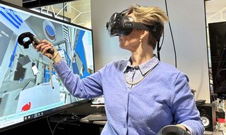 A person in a purple sweater using VR goggles sitting next to a TV screen depicting what is seen with the goggles.