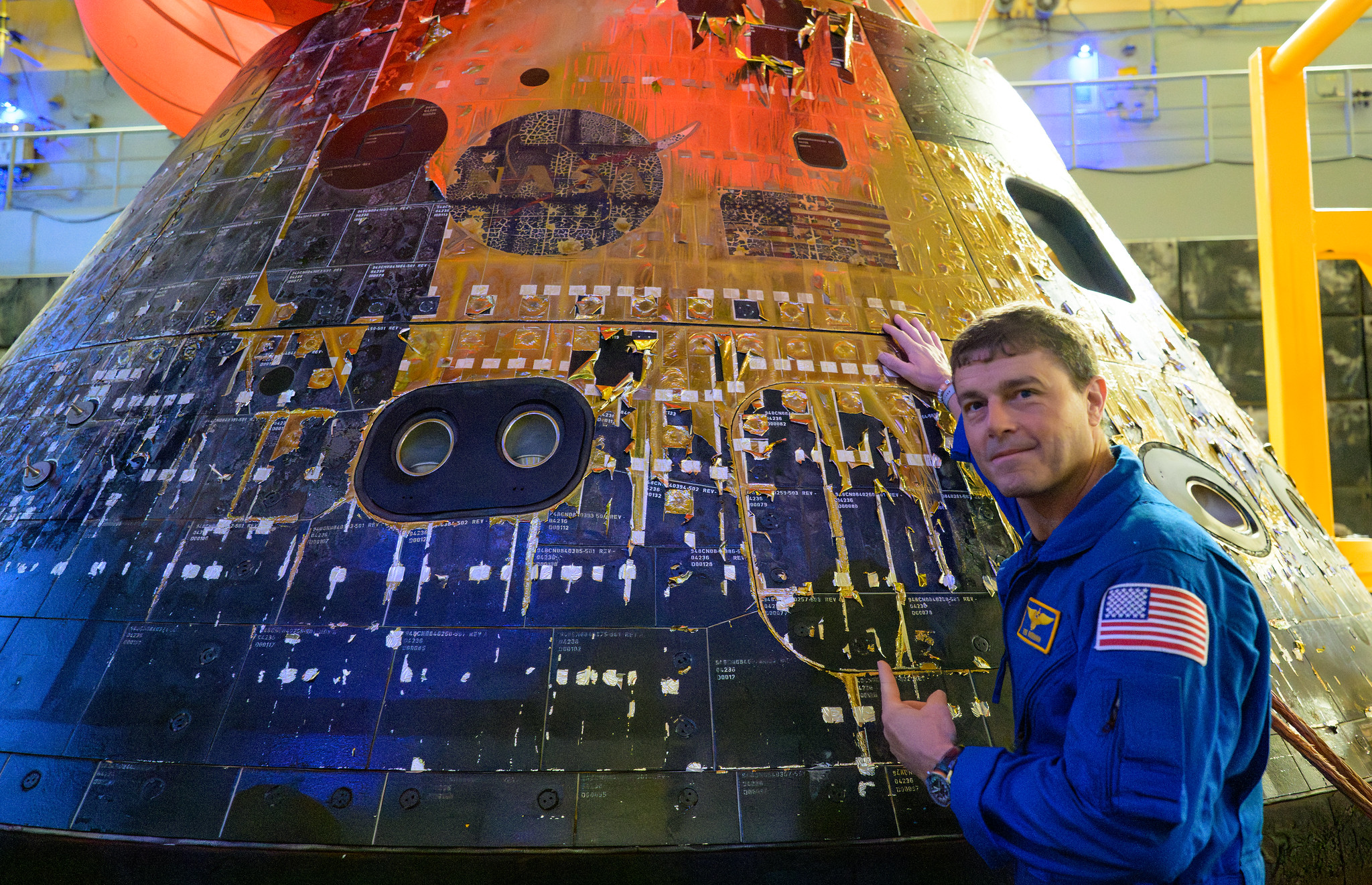 Artemis 2 commander Reid Wiseman is photographed in a blue NASA jumpsuit looking back at the camera with his hand on the charred Orion spacecraft as it sits in the well deck of a recovery ship.