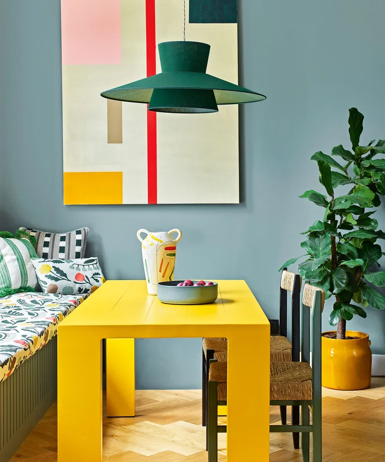 Dining room with blue painted walls, colorful, abstract artwork on walls, large plant in yellow ceramic pot on floor, bright yellow rectangular dining table, built in bench by window, upholstered seat and back with cushions, table decorated with colorful vase and bowl