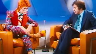 David Bowie with bright orange hair and a colorful costume being interviewed on an old TV in Moonage Daydream