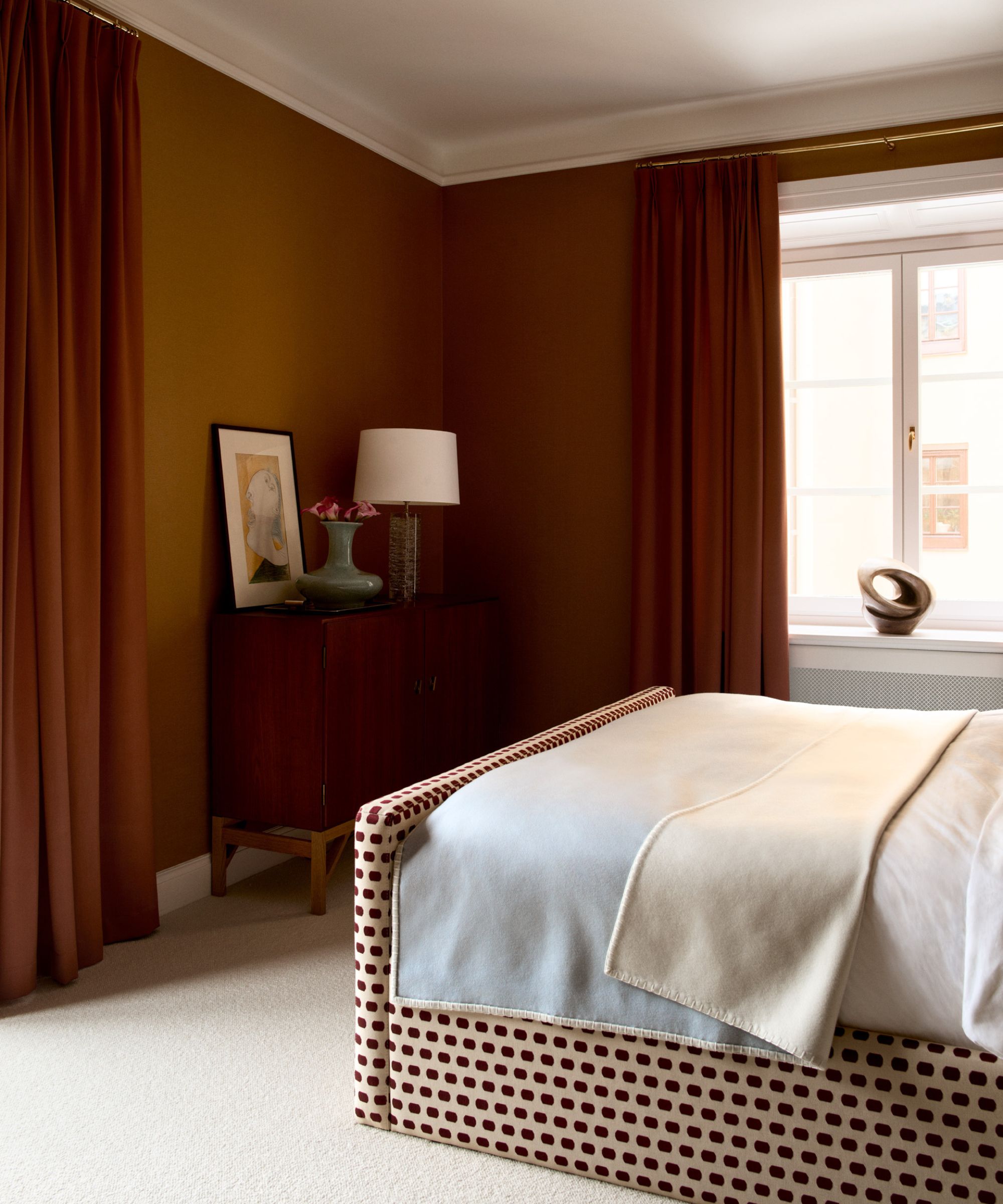 Bedroom with burnt orange walls, an upholstered polka-dot bed with white bedding, two windows with heavy rust-coloured drapes, and a console with a lamp, vase and print propped on top.