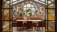 A veranda-style dining room with mural walls, stained glass doors, a pale wood dining table with leather chairs and a brown leather soft bench with green topper, and a thriving plant set in a vase.