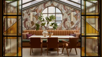 A veranda-style dining room with mural walls, stained glass doors, a pale wood dining table with leather chairs and a brown leather soft bench with green topper, and a thriving plant set in a vase.