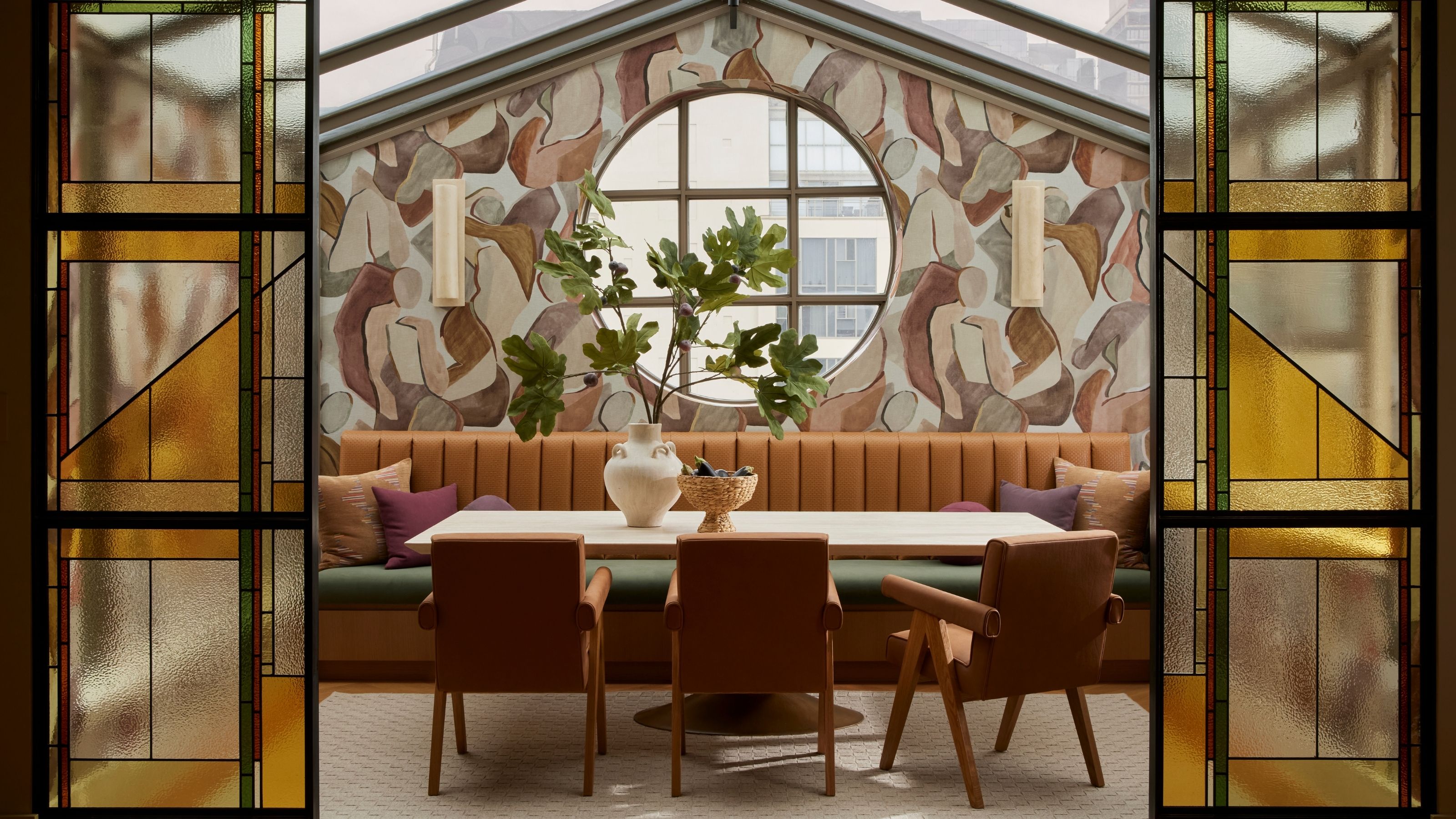 A veranda-style dining room with mural walls, stained glass doors, a pale wood dining table with leather chairs and a brown leather soft bench with green topper, and a thriving plant set in a vase.