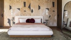 Bedroom with low bed in white and pink linen, wall with graphic pattern behind, large floorstanding mirror and pendant lights either side of bed