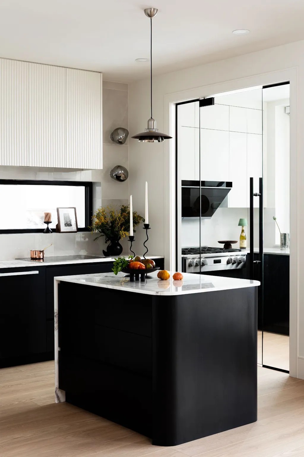 Image of a white kitchen with wooden floors and black, lower cabinetry. There is a small island in the middle painted black with marble countertops. There are two wavy candleholders and fruit on the countertop.